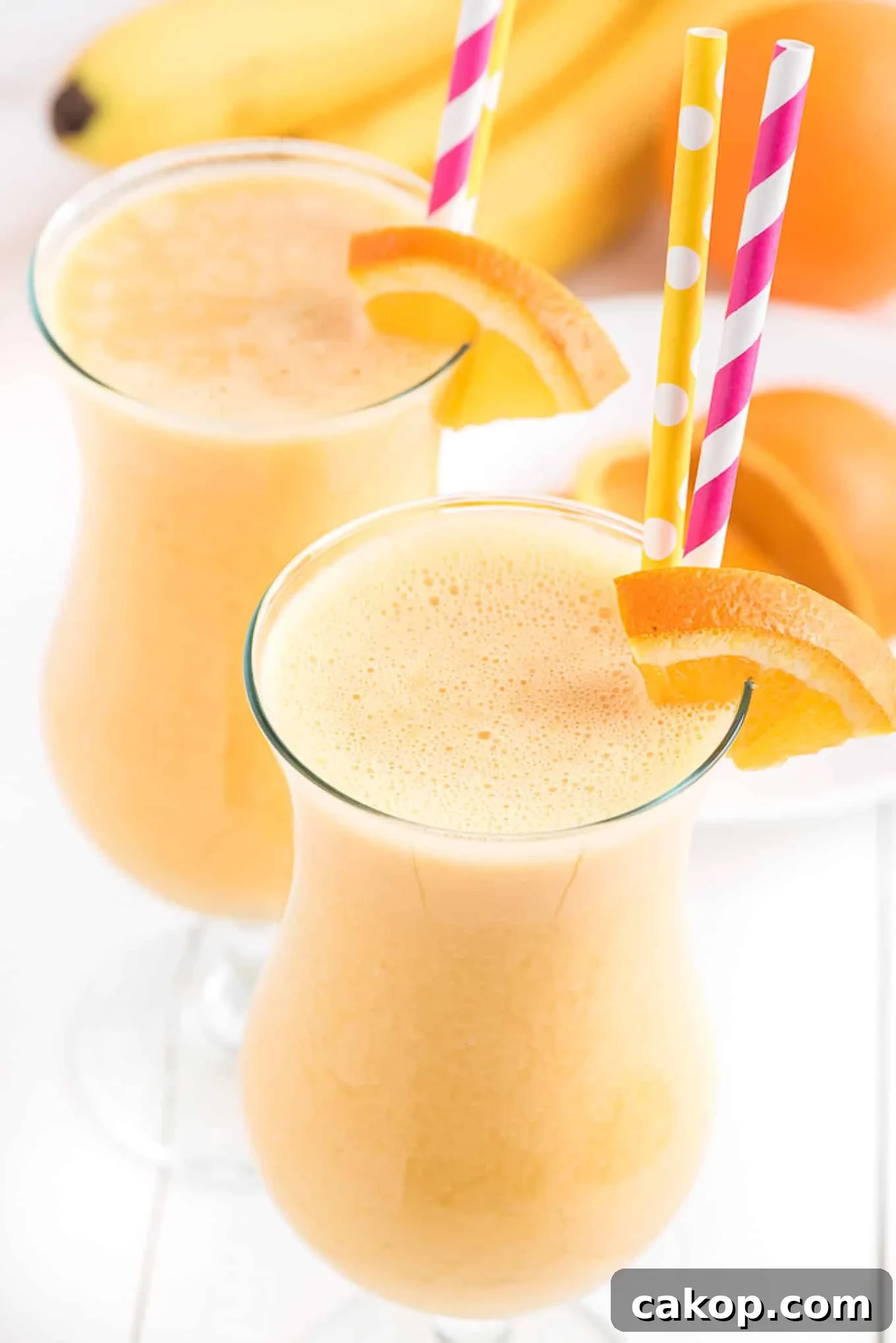 Creamy Orange Smoothie Recipe for Fresh, Zesty Mornings 2 Tall glass of orange smoothie with an orange slice on the side and a pink straw.