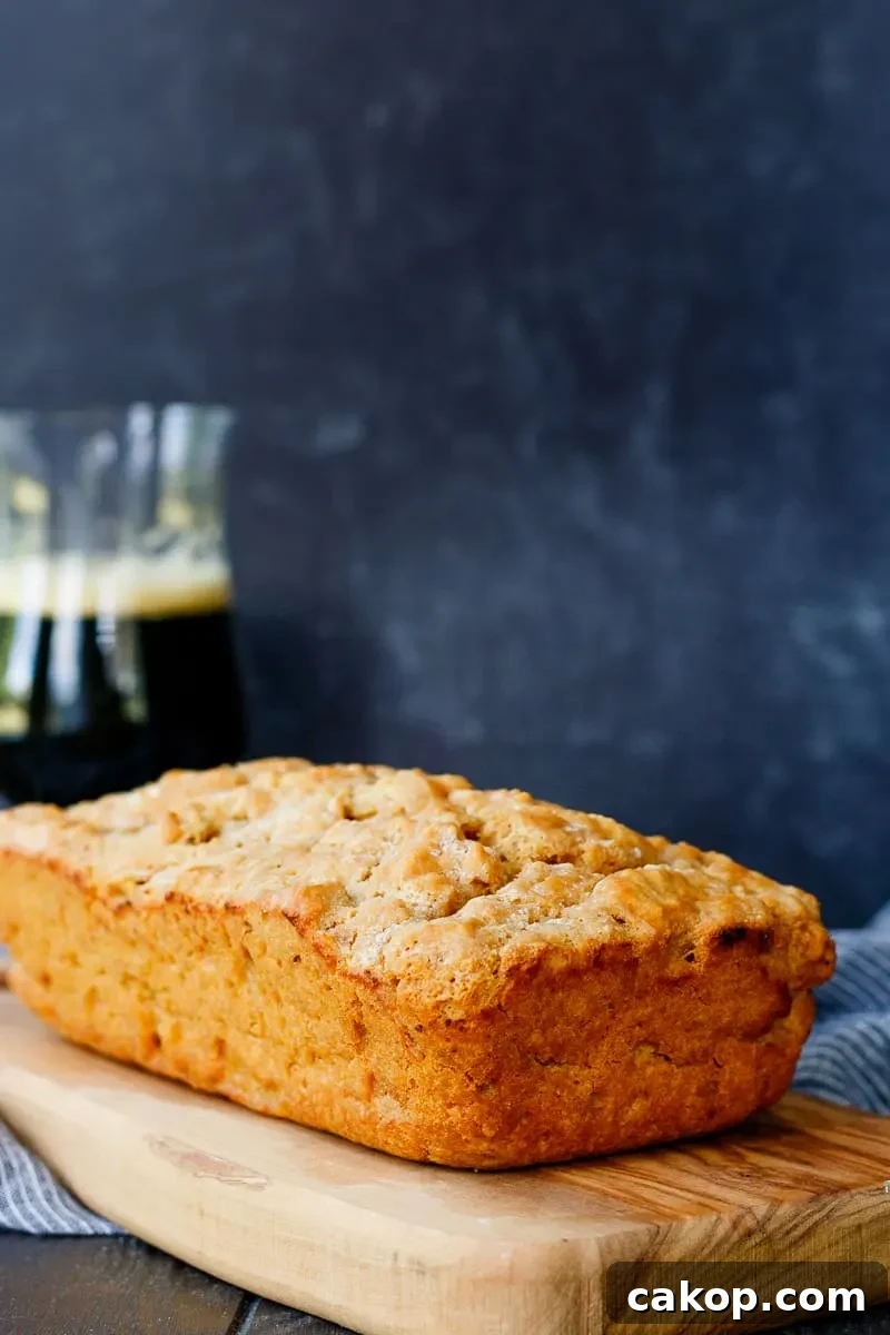 loaf of beer bread on a cutting board with a darker background and glass of beer towards the rear left of the photo