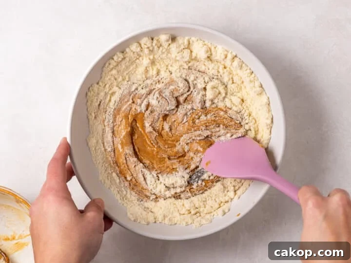 High-Protein Snickers Date Bark Recipe for Satisfying Snacks 11 A pink spatula stirring peanut butter, protein powder, vanilla bean paste, and pure maple syrup together in a mixing bowl