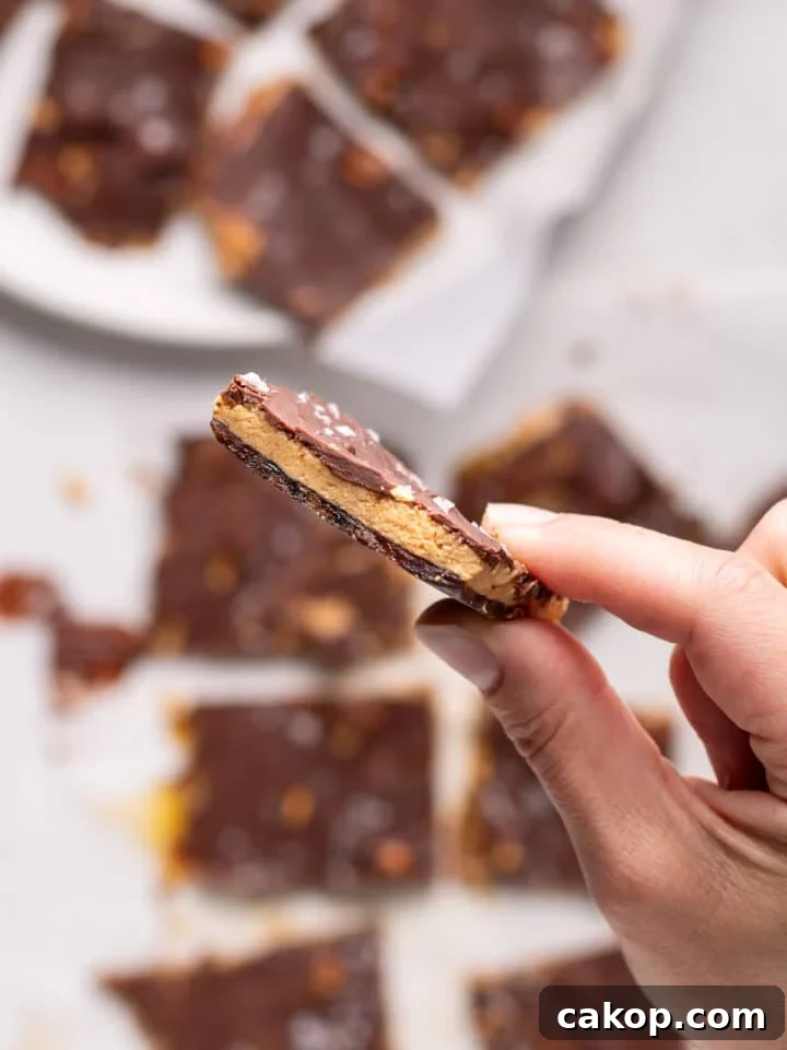 High-Protein Snickers Date Bark Recipe for Satisfying Snacks 19 A hand holding a thin piece of peanut butter date bark with chocolate, peanuts, and flaky sea salt
