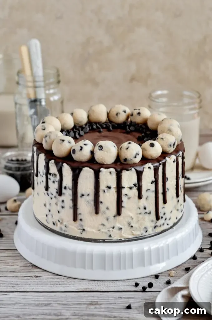 A cookie dough cake covered in cream cheese cookie dough frosting with dark brown chocolate ganache drip and topped with edible cookie dough balls.