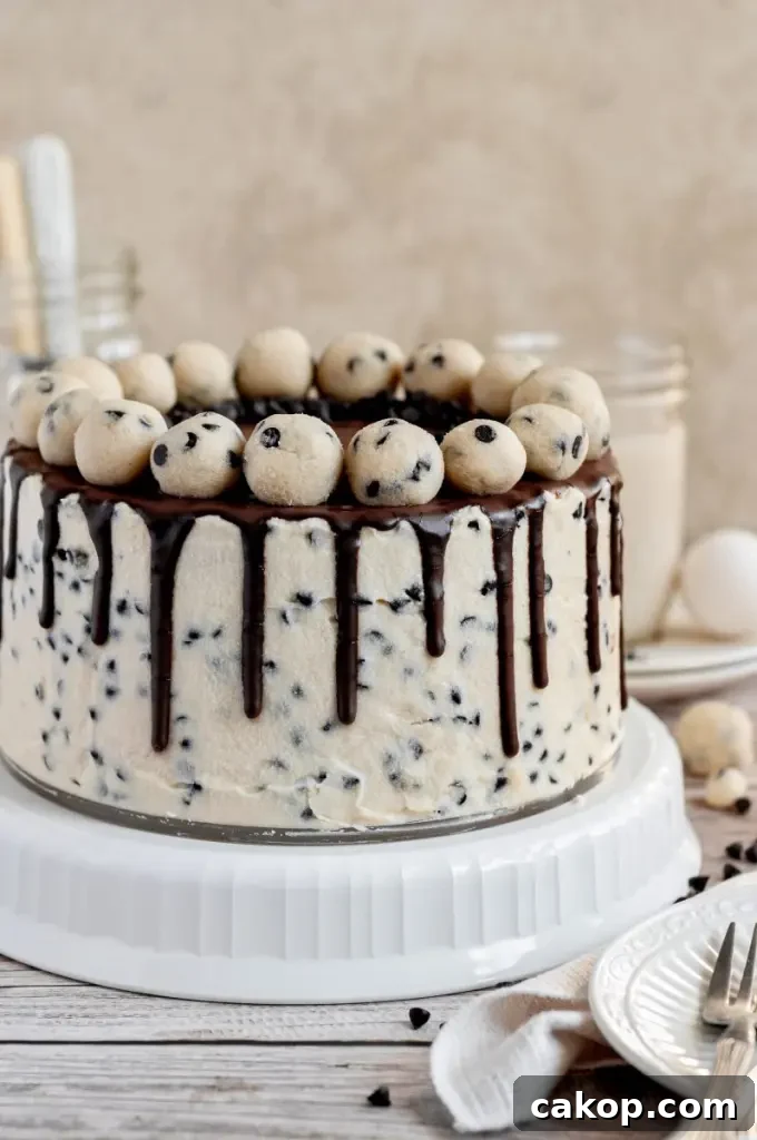 A cookie dough cake covered in cream cheese cookie dough frosting with dark brown chocolate ganache drip and topped with edible cookie dough balls.