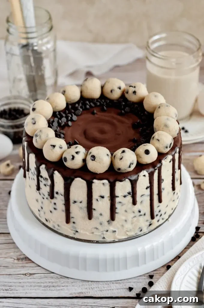 A cookie dough cake covered in cream cheese cookie dough frosting with dark brown chocolate ganache drip and topped with edible cookie dough balls.