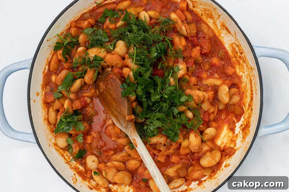 Greek Gigantes Plaki with Canned Giant Beans: Authentic Recipe 6 Overhead of a blue cast iron pan on a marble background with cooked Greek beans in tomato sauce and a wooden spoon and with fresh dill and parsley on top ready to be stirred in.