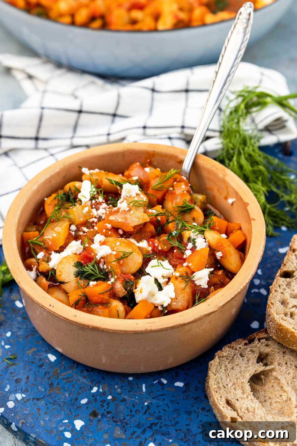 Greek Gigantes Plaki with Canned Giant Beans: Authentic Recipe 7 A ceramic dish of Greek giant beans with fresh herbs and feta crumbled on top, on a blue marble plate stand with slices of bread, fresh dill, a white and blue checked tea towel and a bigger pan of the beans around it.