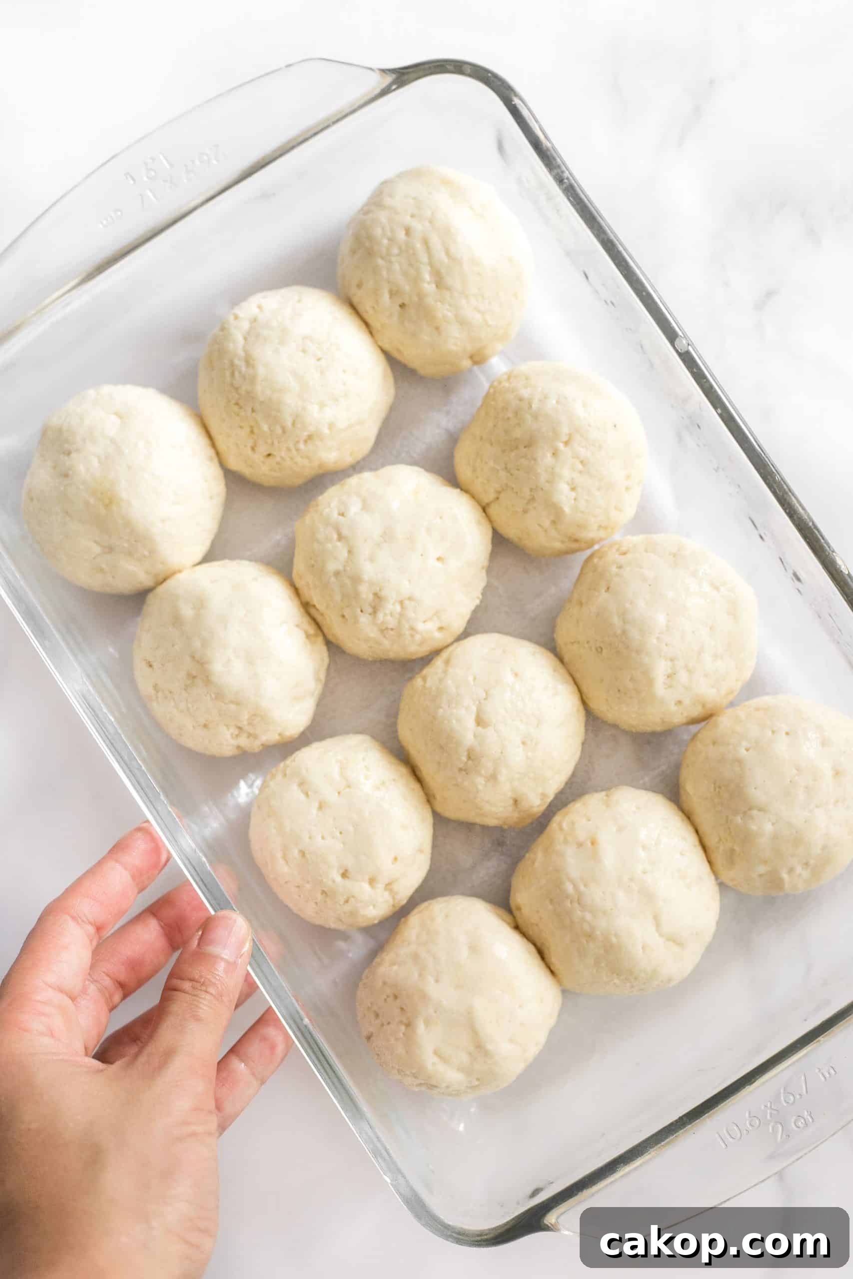 Soft Gluten-Free Dinner Rolls (Dairy-Free) Hand holding a glass pan of balls of bread dough.