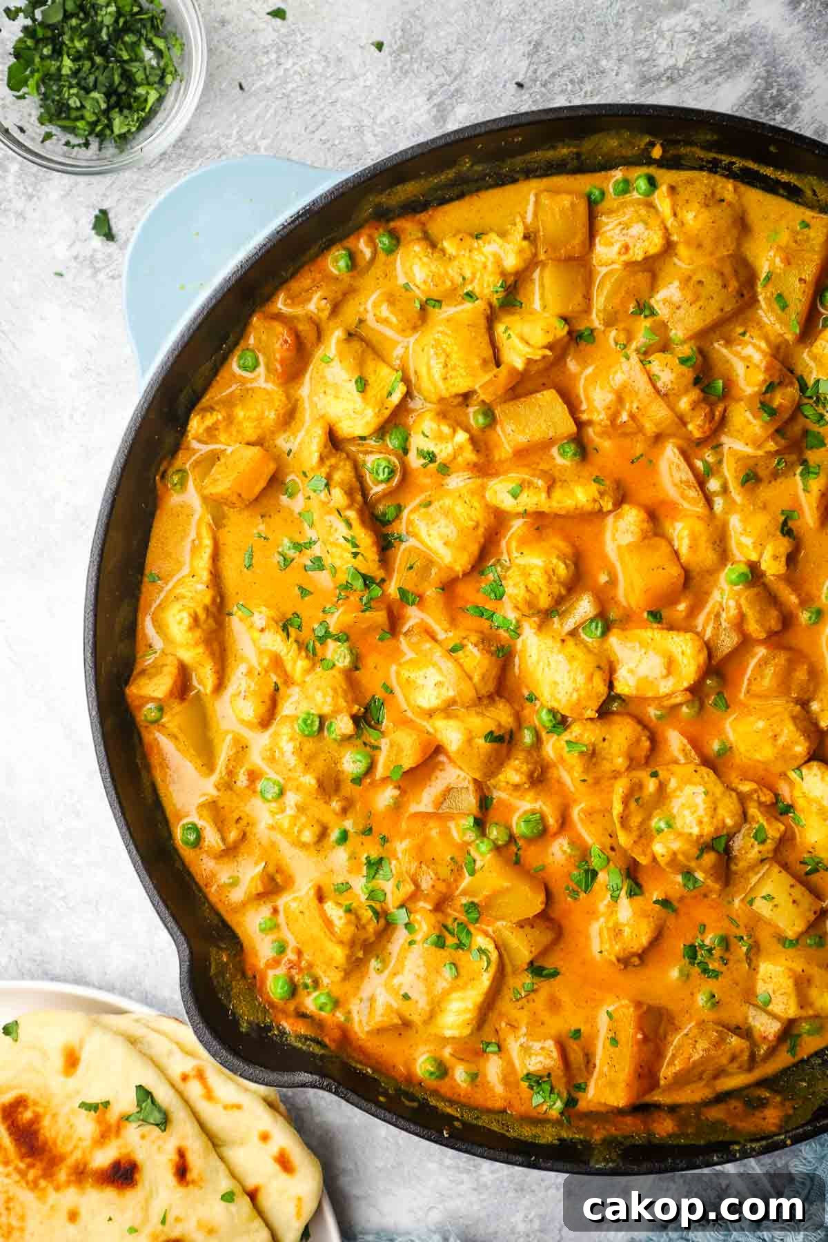A cast iron pan full of chicken and potato curry garnished with cilantro, with a plate of naan in the background.