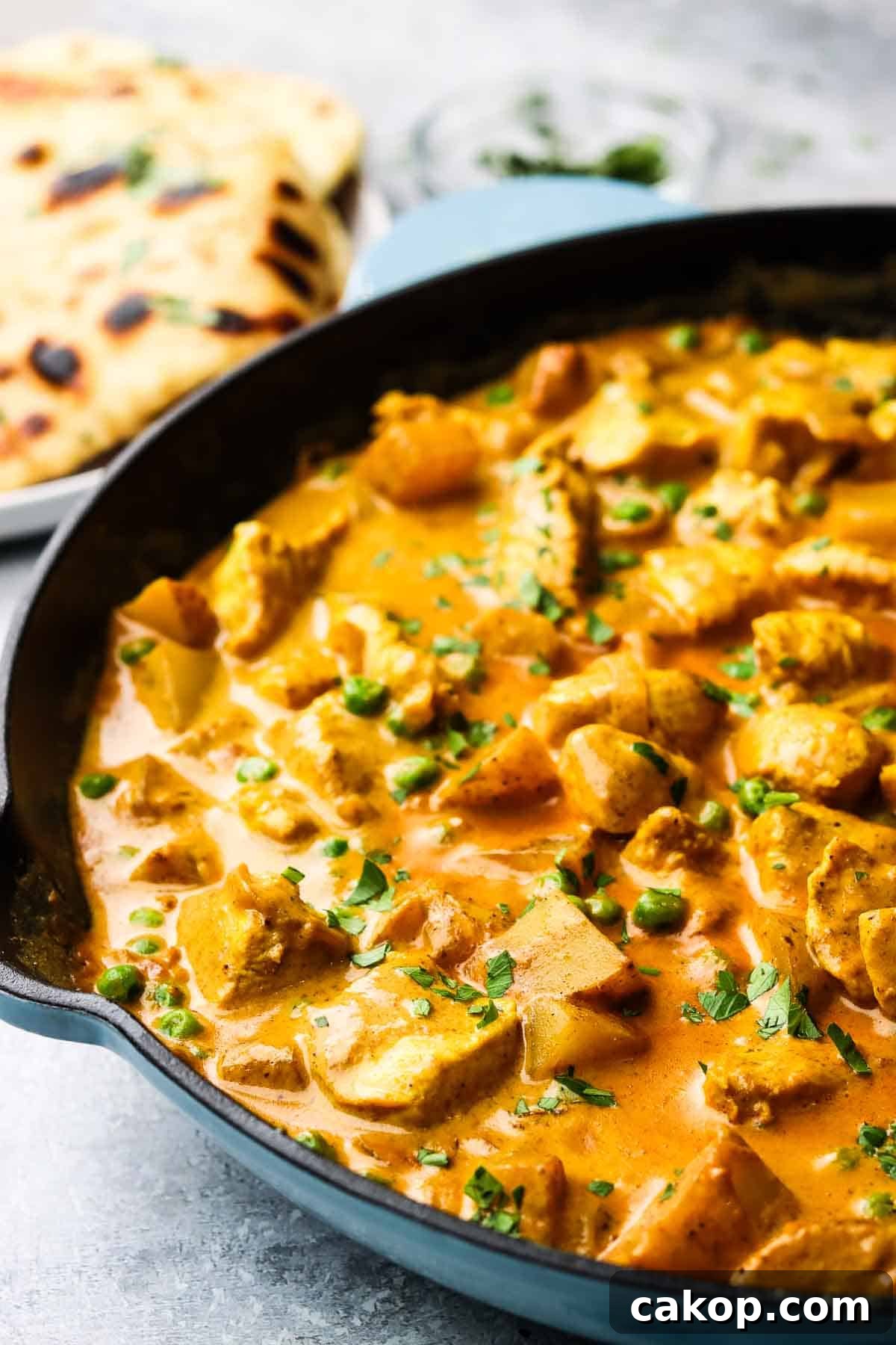 A cast iron pan full of chicken and potato curry garnished with cilantro, with a plate of naan bread in the background.