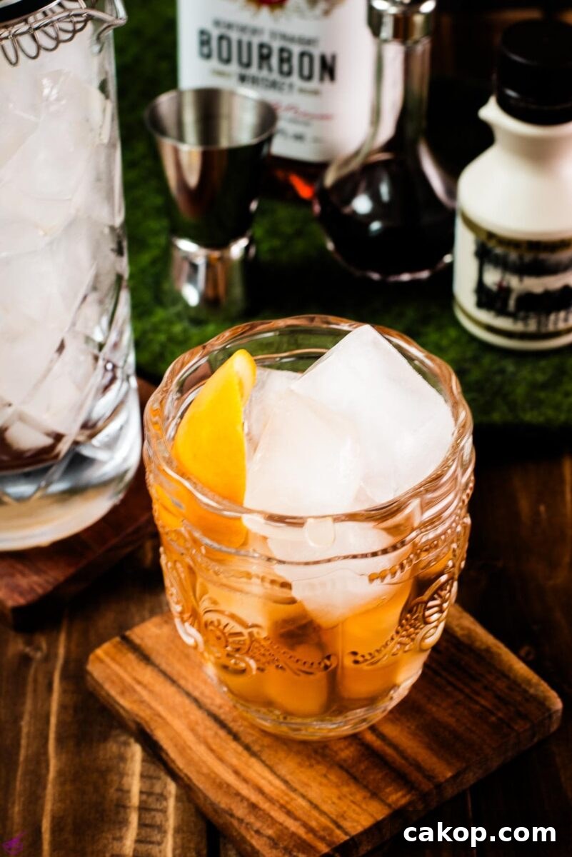 Gorgeous glass filled with maple old fashioned on wooden coaster. The drink is garnished with an orange zest and placed next to a stirring glass and a maple syrup bottle.