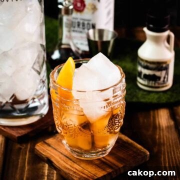 Gorgeous glass filled with maple old fashioned on wooden coaster. The drink is garnished with an orange zest and placed next to a stirring glass and a maple syrup bottle.