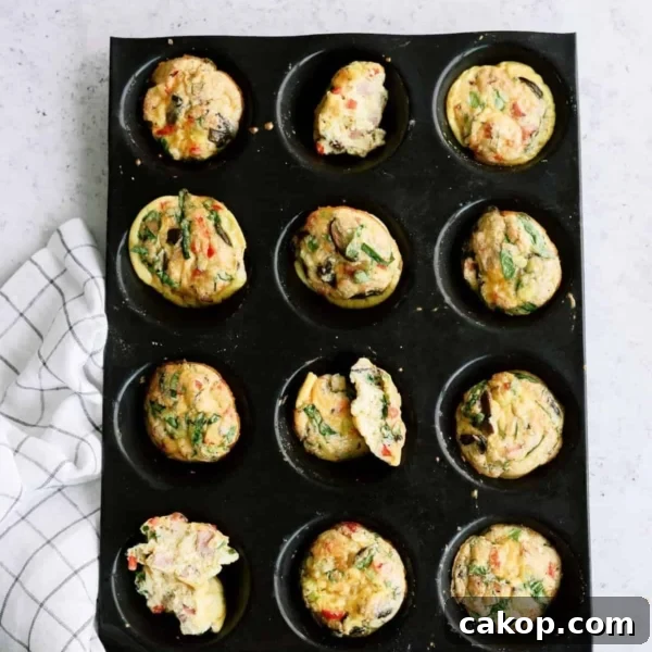 Muffin tray with twelve cooked egg muffins, some with spinach and vegetables, on a light surface next to a checkered cloth.