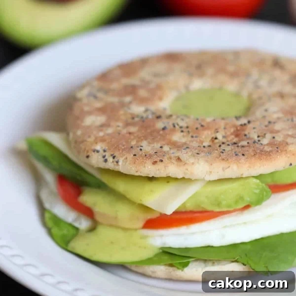 A sandwich on a seeded thin bun with lettuce, sliced tomato, avocado, cheese, and a fried egg. Background shows half an avocado and a tomato.