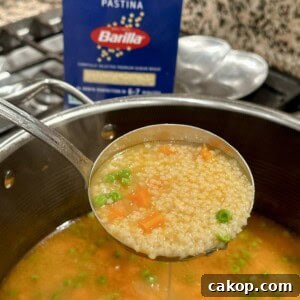 Classic Pastina Soup: Comforting Italian Pasta in a Bowl 5 A ladle holds pastina soup over a pot. The soup contains small pasta, peas, and carrots in a clear broth.