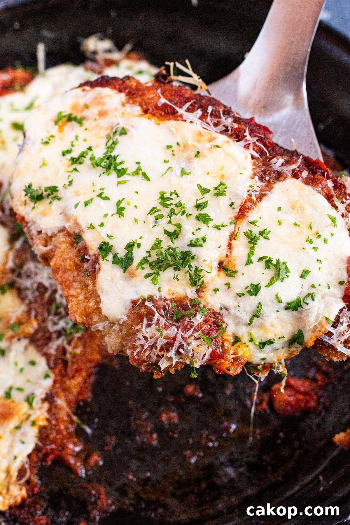 A spatula holding a piece of cheesy chicken parmesan out of a cast iron skillet.