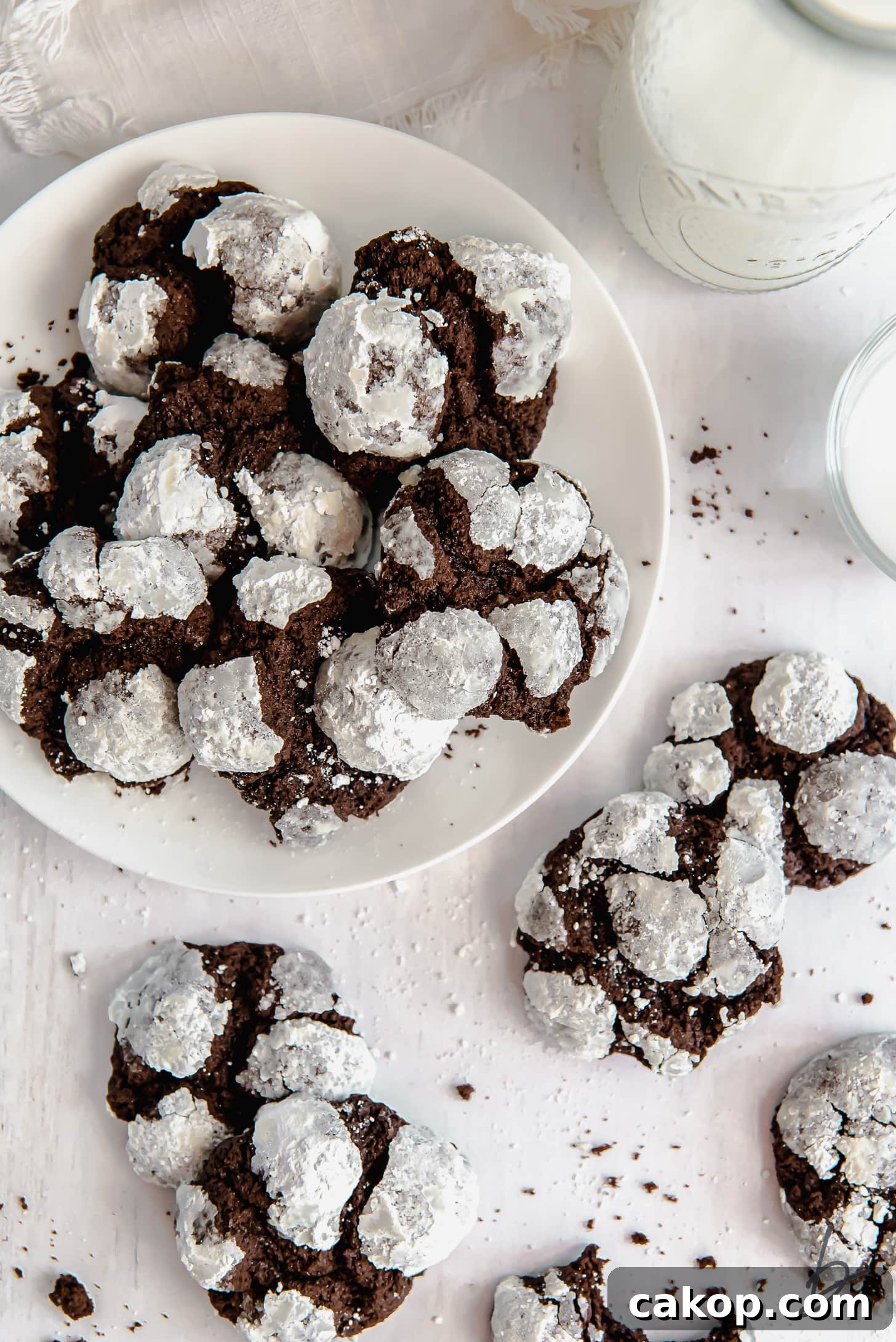 Chewy Chocolate Crinkle Cookie Recipe 2 chocolate crinkle cookies on white round plate next to more cookies and glass of milk