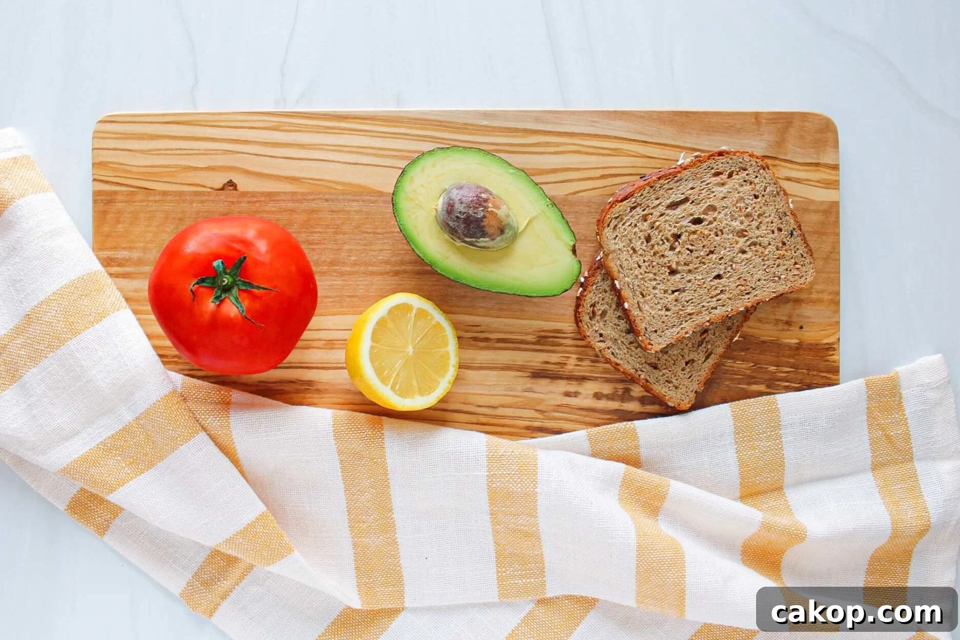 Creamy Avocado Toast with Fresh Tomato and Basil 3 Ingredients for avocado toast with tomato laid out on a wooden board: a tomato, half a lemon, half an avocado, and two slices of whole grain toast.