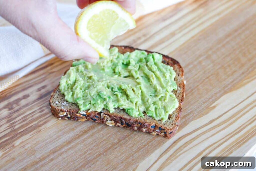 Creamy Avocado Toast with Fresh Tomato and Basil 4 A slice of whole grain bread topped with mashed avocado with a hand squeezing lemon on top.