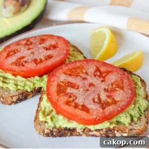 Two slices of avocado toast with tomato on a white plate garnished with lemon slices.