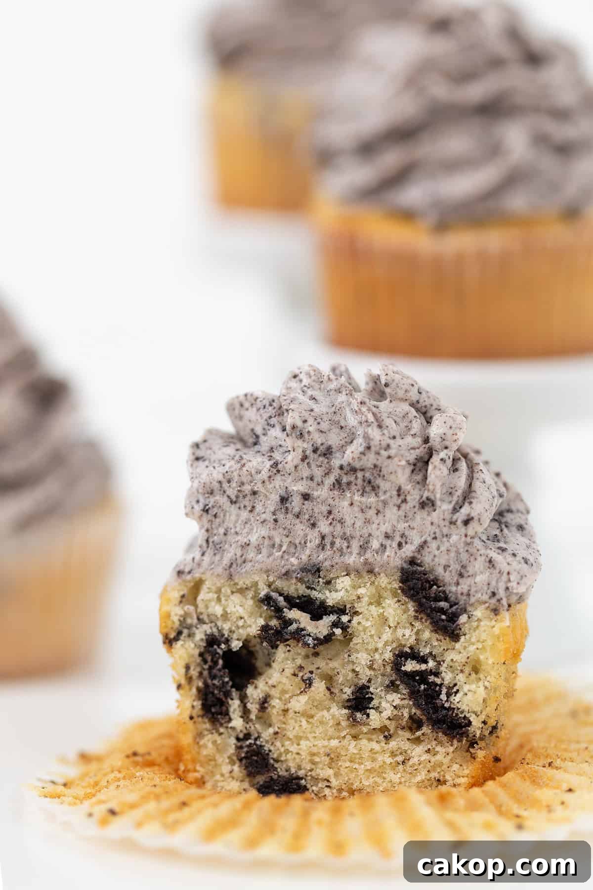 Cookies and Cream Cupcakes Recipe: Rich Oreo-Style Cupcakes 12 A partially eaten Oreo Cookies and Cream Cupcake with cookies and cream frosting sits on an unwrapped liner, revealing chunks of cookie inside. More frosted cupcakes are visible in the background.