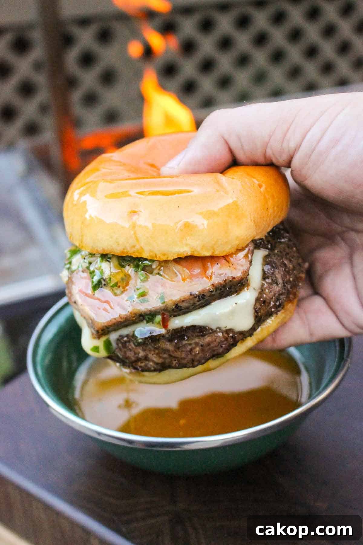 A hand holding a burger over a bowl of cowboy butter for dipping with a live grill fire in the background