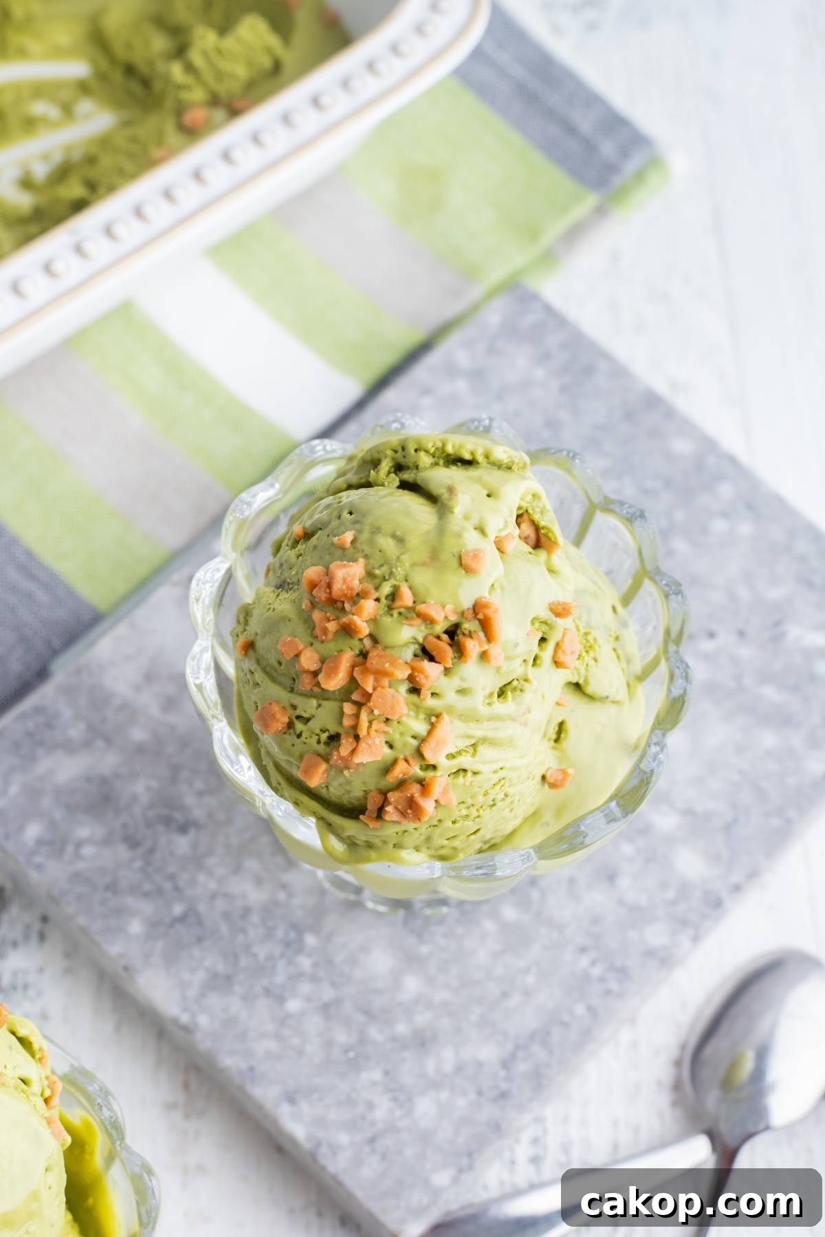 No-Churn Matcha Dulce de Leche Ice Cream Recipe 6 A glass dish of green tea ice cream, made with a no churn ice cream recipe, topped with crunchy bits on a gray stone slab, with a spoon nearby.