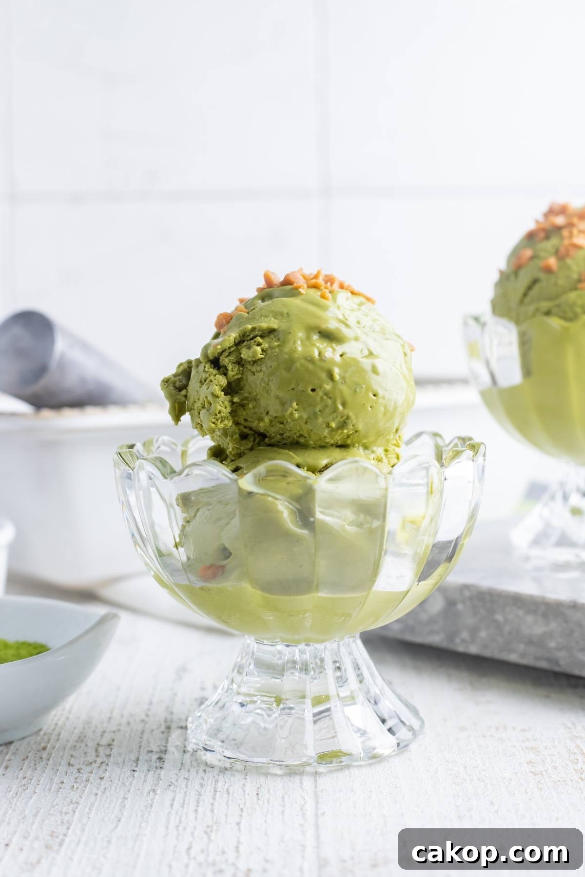 No-Churn Matcha Dulce de Leche Ice Cream Recipe 8 A glass dish with two scoops of green tea ice cream, topped with chopped nuts, on a white table—a perfect treat to try with a matcha ice cream recipe without a machine.
