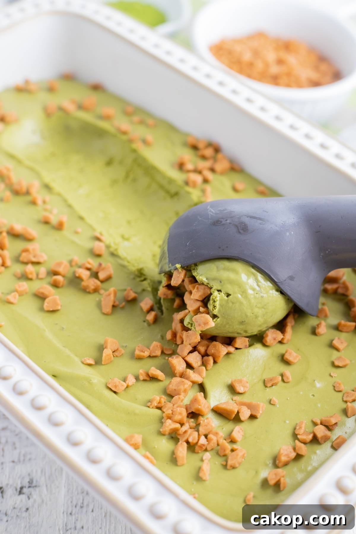 No-Churn Matcha Dulce de Leche Ice Cream Recipe 10 A scoop of matcha ice cream with toffee bits in a white dish, served alongside a gray ice cream scooper—perfect for anyone looking for an easy matcha ice cream recipe without a machine.