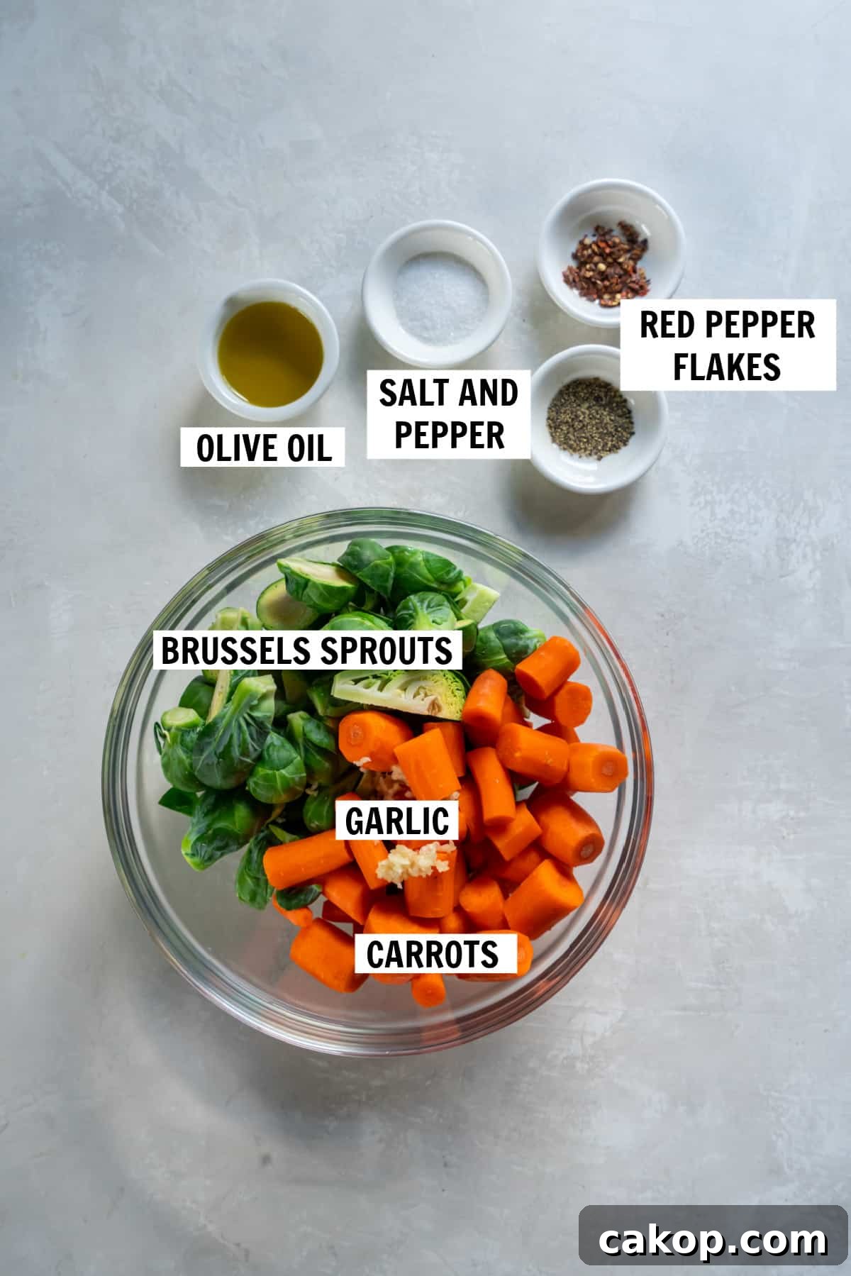 All of the ingredients for roasted brussels sprouts and carrots in bowls on a white countertop including fresh brussels sprouts, fresh cut carrots, minced garlic, olive oil, sea salt, black pepper and red pepper flakes.