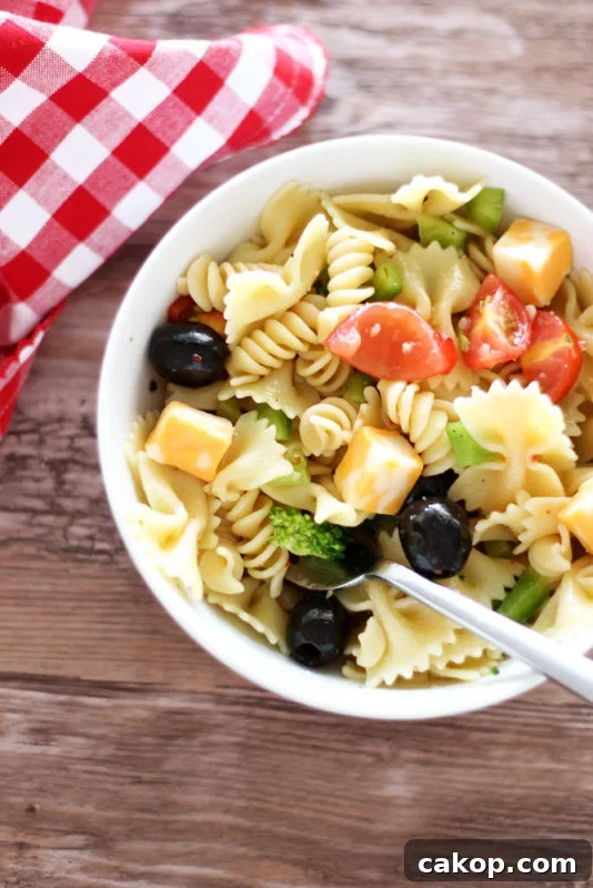 White bowl containing pasta salad topped with tomatoes, peppers, cheese and broccoli, fork in bowl with red gingham napkin on brown table.