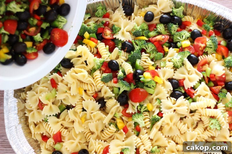Silver pan containing Italian Style pasta salad with tomatoes, peppers, cheese and broccoli being poured in from a white bowl.