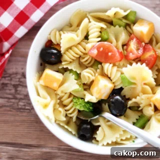 White bowl containing pasta salad topped with tomatoes, peppers, cheese and broccoli, fork in bowl with red gingham napkin on brown table.