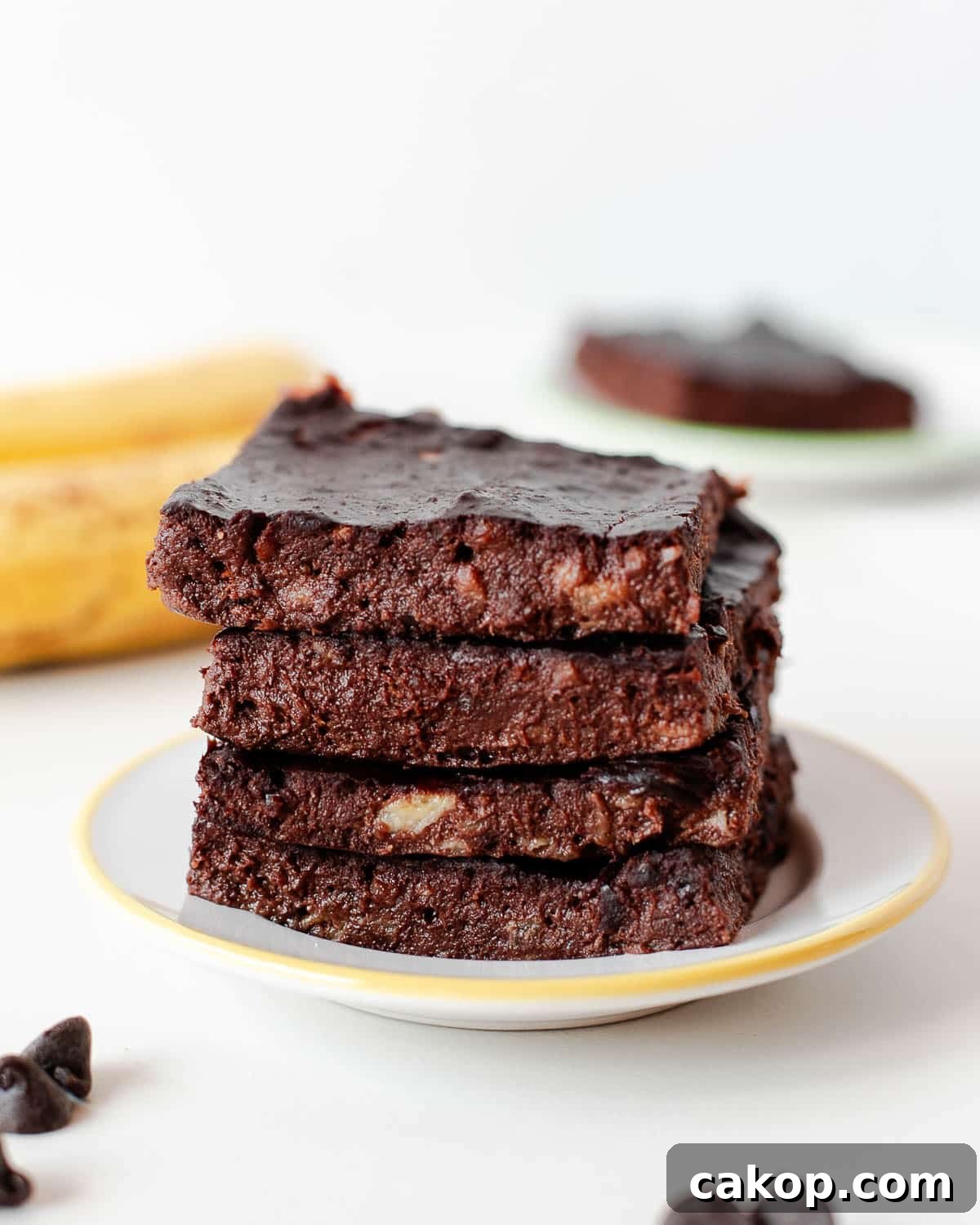 Stack of fudgy 3 ingredient banana brownies on a small plate with chocolate chips and banana in background.