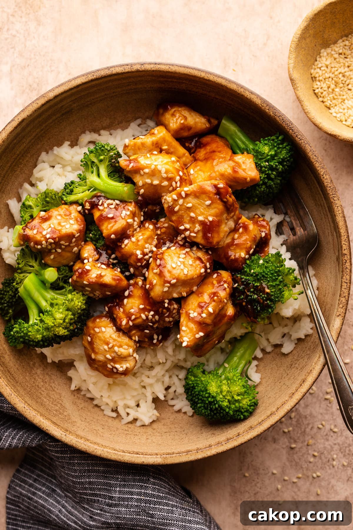Quick Teriyaki Chicken Stir-Fry Ready in 10 Minutes 2 A tan colored bowl with teriyaki chicken and broccoli served over rice. The bowl is sitting on a tan background with a small bowl of sesame seeds and a dark napkin arranged around it.