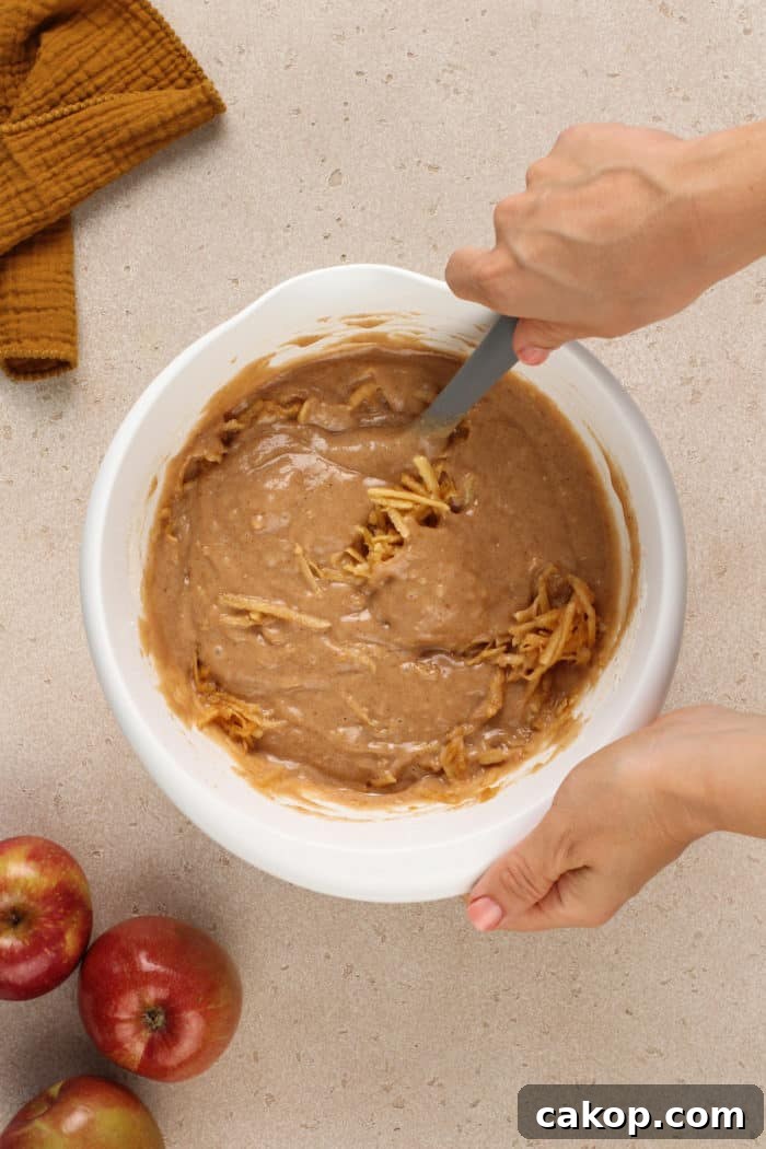 Grated apple being folded into apple butter cake batter in a white mixing bowl.