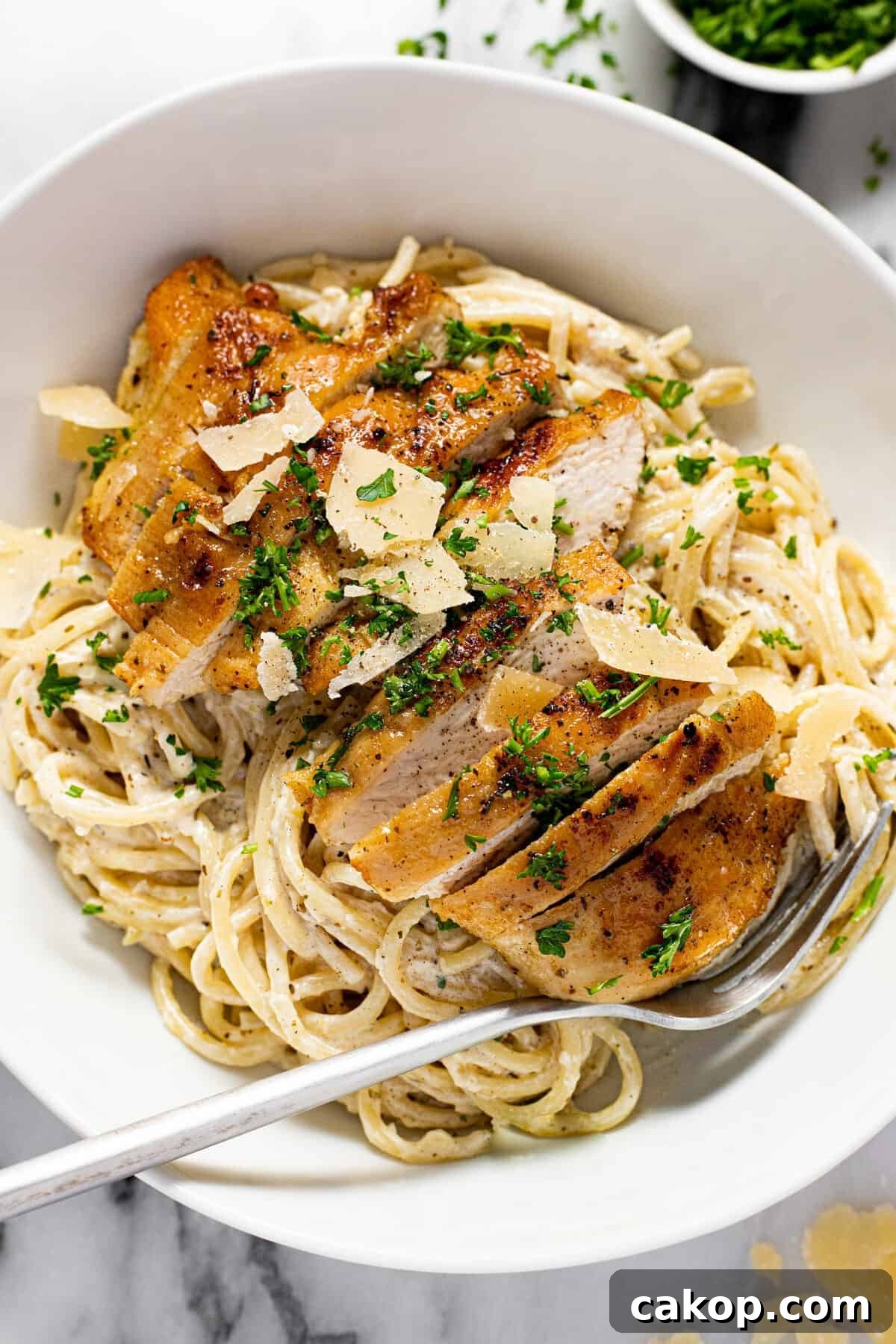 Large white bowl filled with creamy spaghetti and sliced lemon chicken garnished with parsley.
