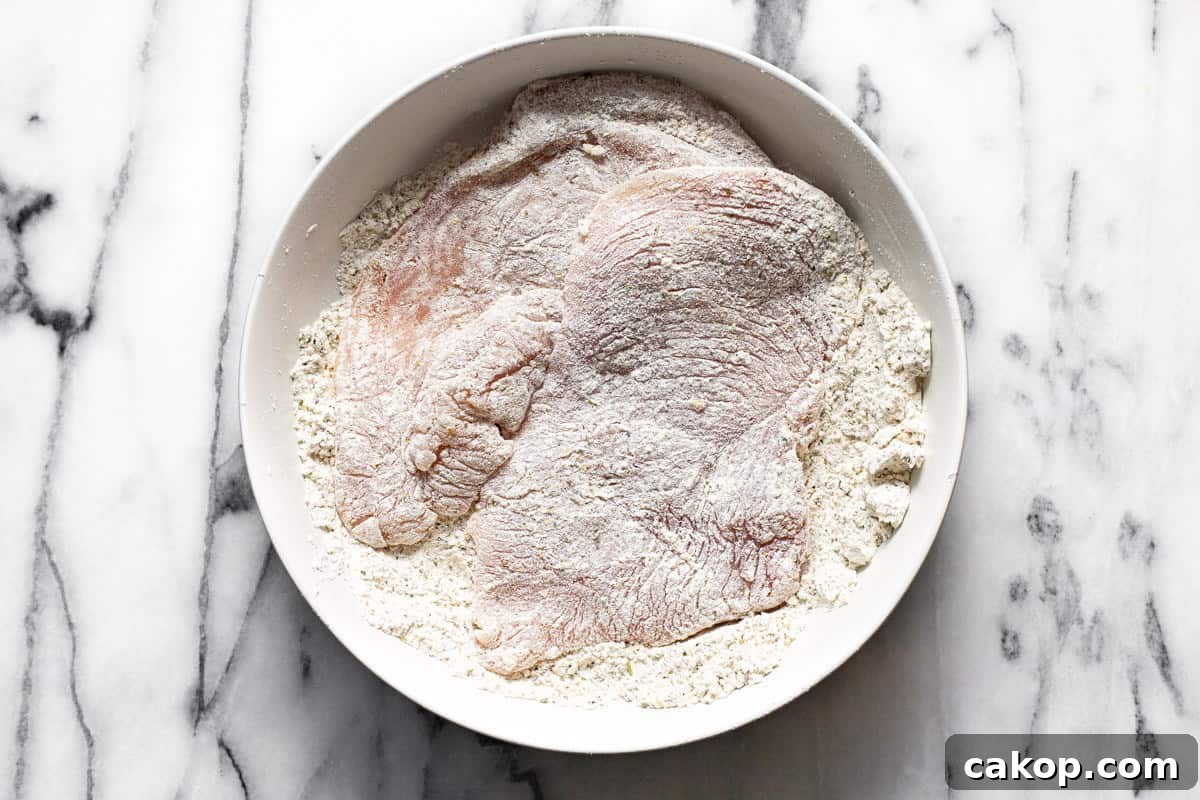 Chicken breast dredged in a white bowl filled with flour and seasonings.