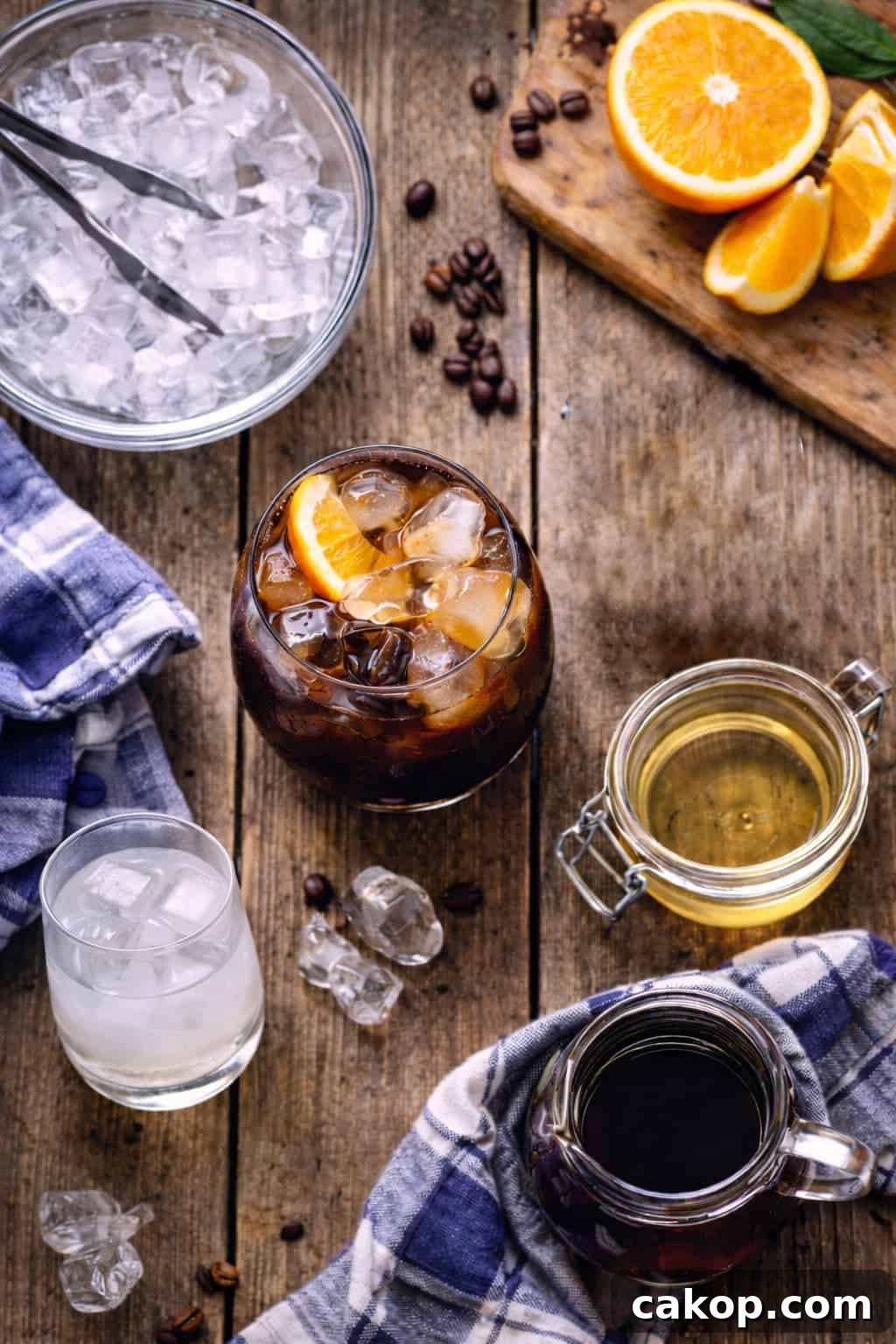 Ingredients for ouzo cocktail including coffee liqueur, orange slices, fennel syrup and ice on a rustic wooden surface.