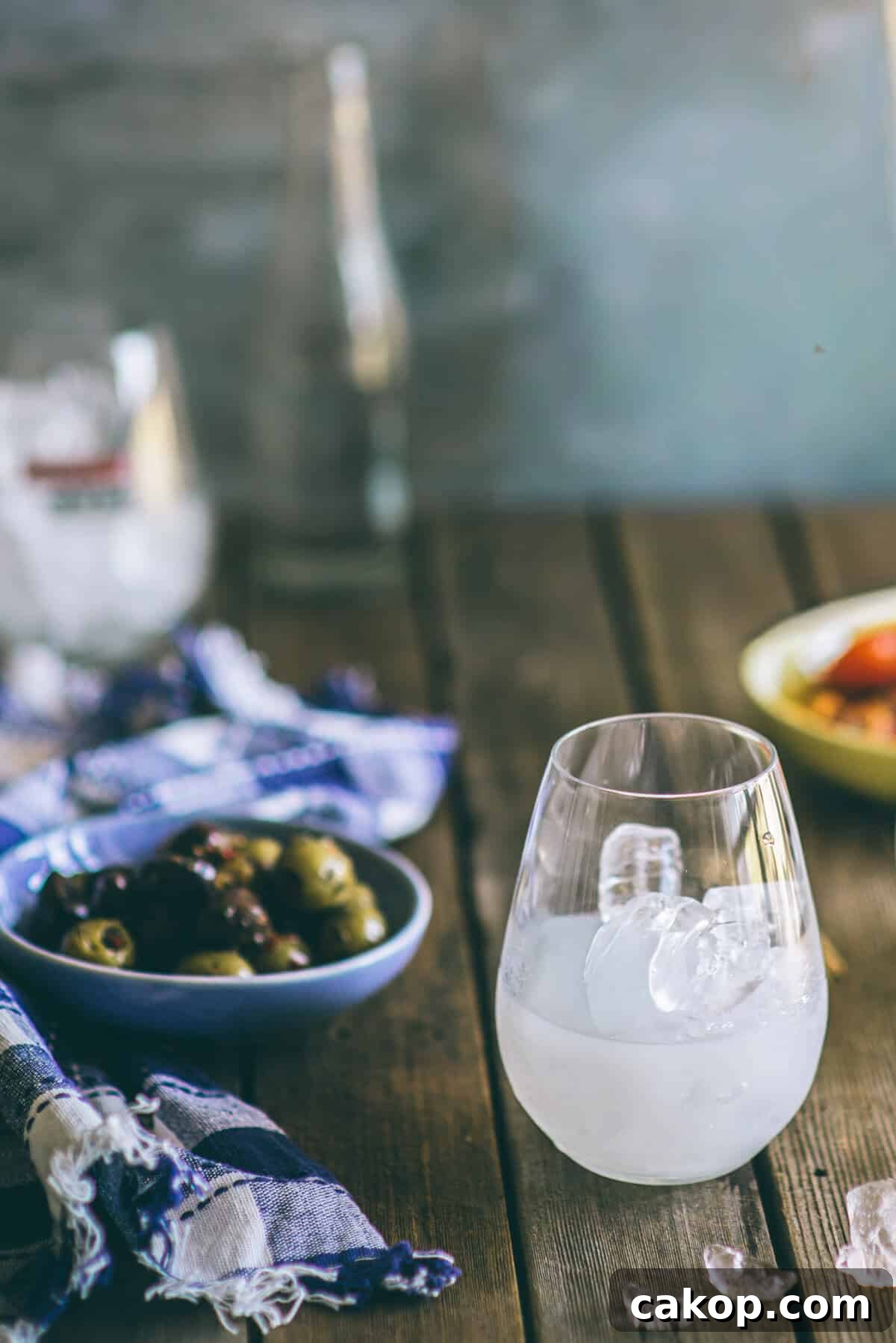 Glass of ouzo with ice turning cloudy alongside olives on a rustic table setting.