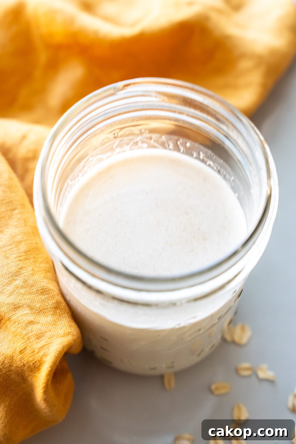 Creamy Homemade Oat Milk Recipe You Can Make at Home 2 A glass jar filled with creamy white liquid, placed on a light surface with a few scattered oats nearby. A yellow cloth is partially visible to the left.