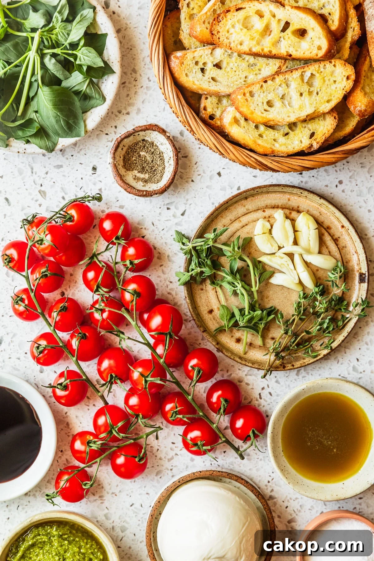 White and brown bowls of garlic, herbs, oil, vinegar, crackers, basil, mozzarella, and pesto on a white terrazzo counter next to tomatoes.