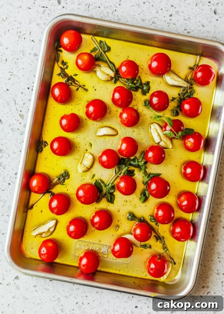 A silver sheet pan of cherry tomatoes tossed with oil, garlic, and herbs on a white counter.