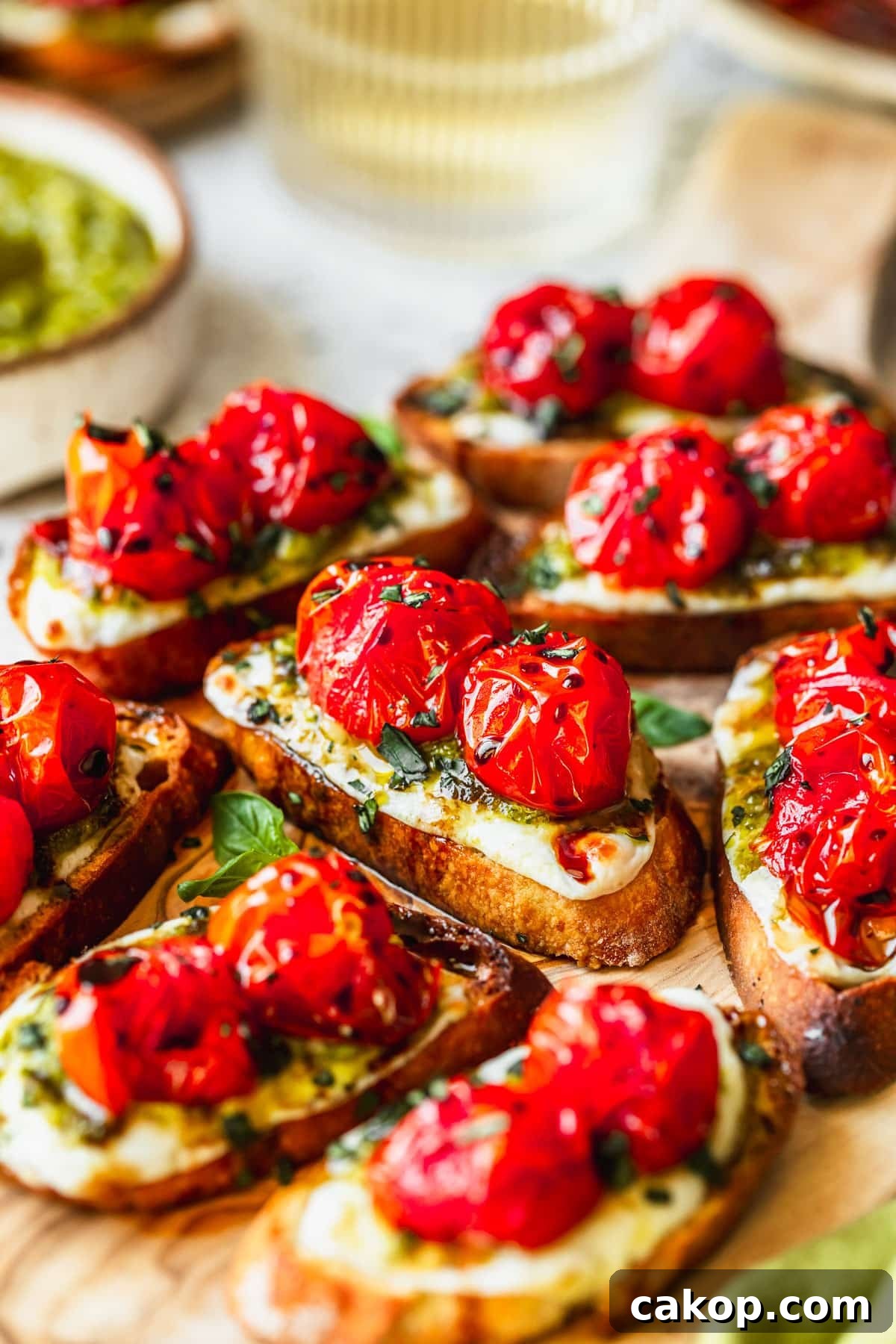 A wood board of Caprese crostini on a white counter next to glasses of white wine and a white bowl of pesto.