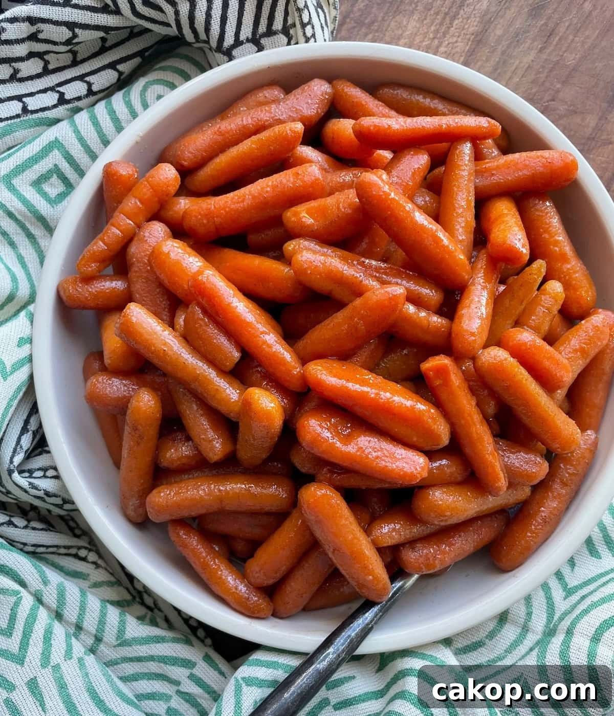 A bowl of shiny glazed baby carrots with a spoon, placed on a patterned cloth, viewed from above.