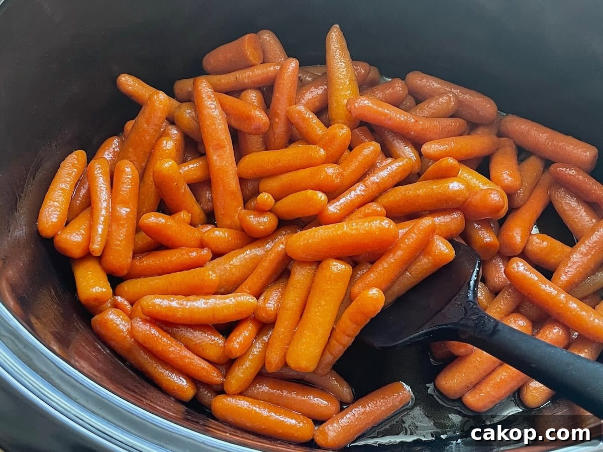 A heap of shiny glazed baby carrots cooked in a slow cooker with a black serving spoon.