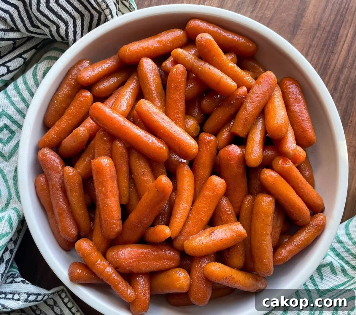 A white bowl filled with shiny glazed baby carrots on a wooden table, with a patterned cloth napkin alongside.