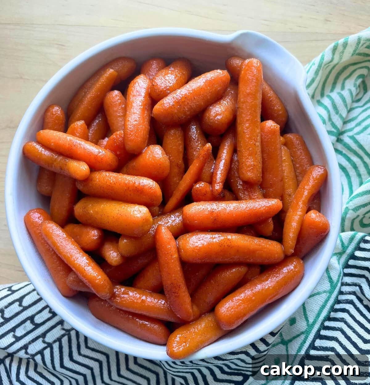 A white bowl full of shiny glazed baby carrots on a wooden surface with a green and white cloth beside it.