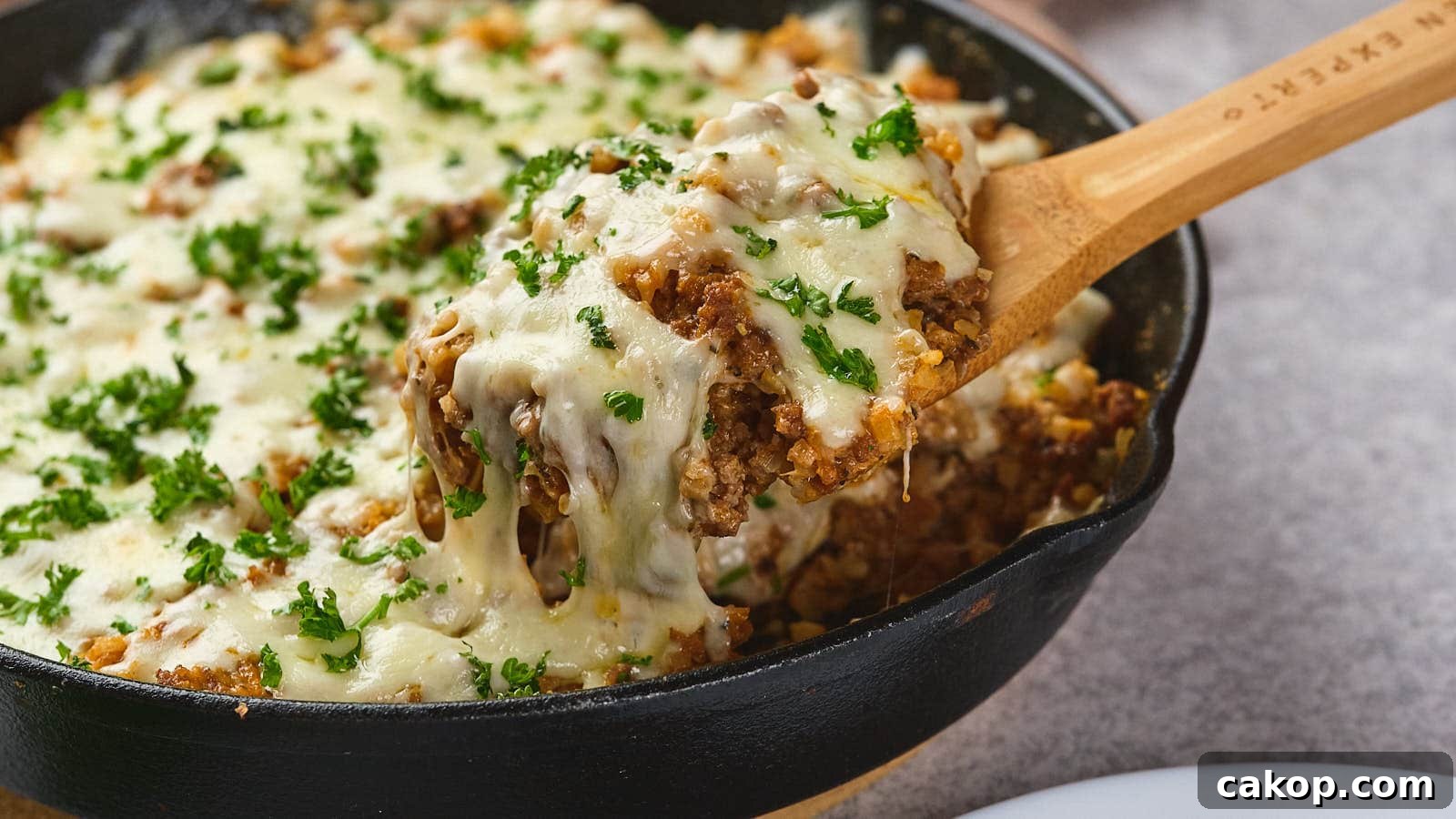 A wooden spoon lifting a serving of italian ground beef skillet garnished with parsley from a cast-iron skillet.
