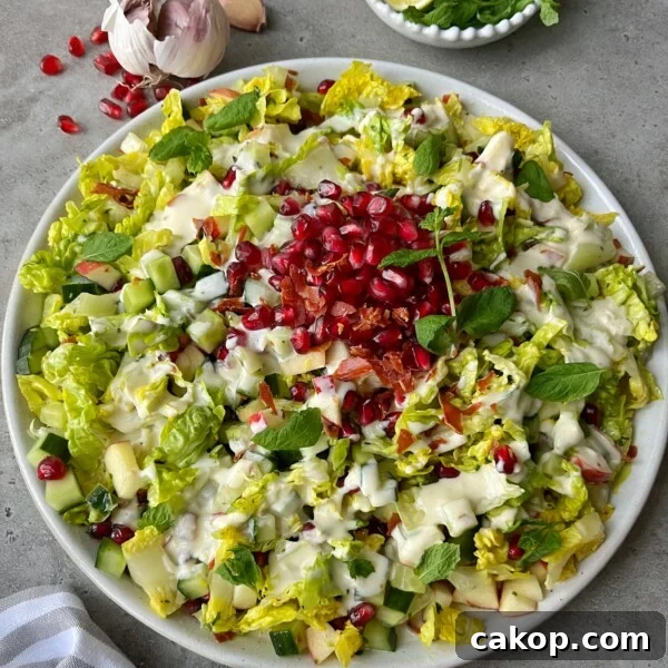 A plate of Crispy Prosciutto Salad topped with pomegranate seeds, chopped cucumber, diced apple, fresh mint leaves, and creamy dressing. Garlic, mint, and a striped napkin surround the plate on a gray surface.