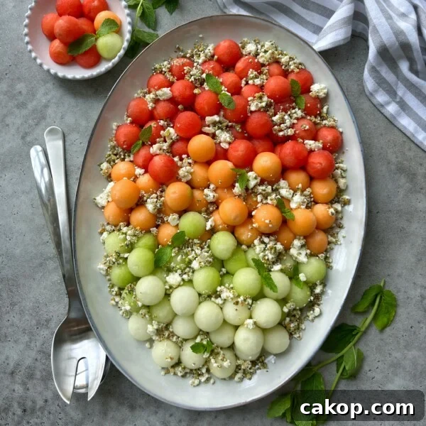 An oval platter of melon feta salad features rows of red, orange, and green melon balls topped with crumbled cheese, herbs, and seeds. Fresh mint leaves, serving utensils, a striped napkin, and a bowl of extra melon balls complete the scene.