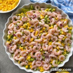 A round dish filled with a vibrant shrimp cocktail salad featuring diced mango, avocado, chopped red onion, and lettuce. Nearby are a bowl of mango cubes and a lemon wedge on a gray surface with a blue striped cloth.